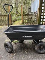 Front-left angled view of black Gorilla Cart yard wagon on outdoor dirt surface in front of wooden fence, showing handle, wheels and plastic bed.