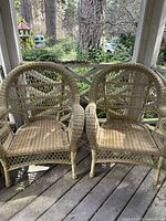 Two light-colored wicker outdoor chairs with rounded backs and armrests, showing woven lattice pattern, placed on a wooden porch.