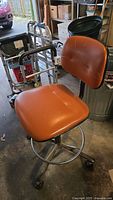 Image showing entire rolling shop chair with orange padded seat and backrest, chrome metal base with wheels, and footrest ring.
