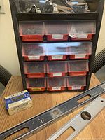 Front view of the black multi-compartment storage organizer with 10 translucent red drawers containing various screws and nails, some labeled. Two metal levels and a boxed hardware item placed in front on a wooden surface.
