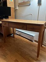 Full view of the wooden desk showing the front side with 3 drawers and tapered legs, placed on hardwood flooring against a wall with a heater below.