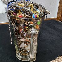 Front side view of the glass container showing the handle and assorted tangled jewelry inside, including silver and gold tone chains, colorful beads and bracelets.