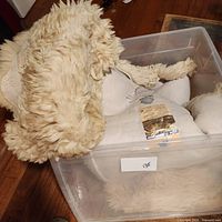 Two cream/off-white Australian lambskin rugs shown in a plastic container on wooden floor, one partially draped showing wool texture.