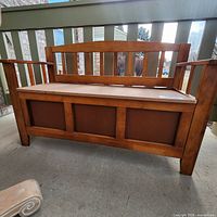 Front view of solid wood bench with slatted backrest and arms, flat seat with lift-up storage compartment underneath.