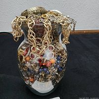 Front view of clear glass vase filled to the brim with assorted costume jewelry including chains, colorful bead necklaces, and metallic pendants showing condition and detail