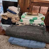 Photo showing neatly folded queen-sized sheets in various colors stacked along with assorted blankets including a white flannel blanket with green print and thick wool and textured blankets.