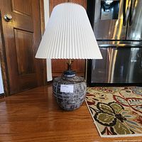 Full view of vintage table lamp with ceramic patterned base and pleated white shade on hardwood floor in room.