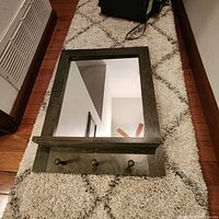 Top-down photo of vintage mirror with wood frame, shelf, and three brass coat hooks placed on a patterned carpet floor.