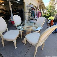 Vintage dining set sitting outdoors on concrete with round glass top table and four cream upholstered chairs showing upholstery and wooden frames.