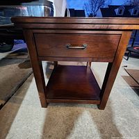 Front view of brown wooden nightstand with drawer and lower shelf on concrete floor at dusk.