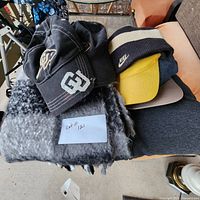 Collection of hats on top of a gray and black scarf with folded jackets and hoodies beneath.