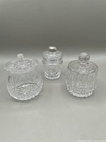 Three clear crystal jars with lids of varying shapes and intricate cut designs shown from front view.