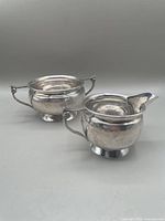 Silver plate cream jug and sugar bowl with hand hammered texture and Arts and Crafts style design, placed on a grey surface.