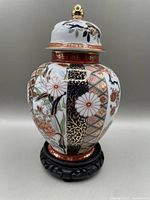 Front view of oval-shaped Japanese ginger jar decorated with Imari floral and geometric patterns in gold, blue, red, and black on white porcelain; topped with lid featuring gold finial, displayed on black carved wooden stand.
