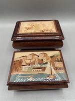 Two wooden musical jewelry boxes; one with light burled wood inset on the lid and the other with an inlaid wood scene of coastal buildings and boats.