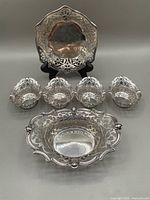 Front view showing 6 silver plated pierced dishes arranged: large bowl, square plate on stand, and 4 small dishes in front.