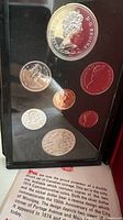 Full display case showing seven coins including the large and small Centennial Winnipeg silver dollars, other standard Canadian coins with Queen Elizabeth II on the obverse, and accompanying description text at bottom.