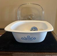 White ceramic Dutch oven with blue cornflower design and glass lid shown separately on a black mat.