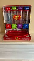 Front view of Jelly Belly My Favorites Bean Dispenser in original packaging showing four clear compartments with colored lids and levers and Jelly Belly logo on base.