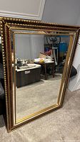 Large vintage mirror with ornate gold and burgundy frame, standing on carpeted floor against wall. Shows full mirror and detailed frame.