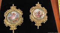 Top-down view of pair of vintage Victorian porcelain wall plaques with gold ornate frames on black background, showing detailed countryside scenes and measuring tape for size reference.
