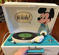 Open view of the record player showing Mickey Mouse inside the lid, turquoise blue turntable base, and Mickey hand stylus arm on the black vinyl record.