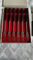 Nine vintage fondue forks with black handles neatly arranged in a box with red felt lining.