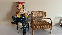 Resin clown figurine seated on metal bench holding playing cards, beside a miniature wicker bench, on a white surface against a plain wall.
