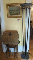Image of farm scene painting on wall with gold frame above wicker basket and nesting tables, floor lamp to right