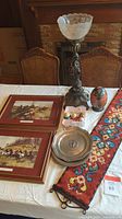 Full table view showing cherub lamp, framed prints, decorative eggs, porcelain egg, pewter plate, knit wall hanging.