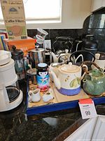 Photo of multiple coffee and tea preparation items including ceramic and metal teapots, French presses, coffee filters, and a coffee maker.