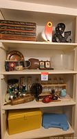 Four shelves displaying assorted items including doll furniture boxes, bird lamp, ampersand sign, decorative plate, cowbell, small picture frames, glass bowl, various vases, musical instruments, figurines, storage cases.