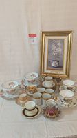 Full group showing original painting with 13 teacup and saucer sets in front on white cloth background