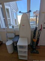 Photo shows collection of household utility items against a window including Simplehuman trash can, white trash can, ironing board, plastic storage drawers and vacuums.