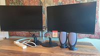 Two Lenovo 27-inch monitors displayed side by side on dual monitor stand with Harman/Kardon speakers and power strip on table