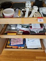 Multiple kitchen and dining items including pewter serving, coasters, a hand mixer, waffles iron and linens shown inside wooden drawers.