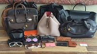 Overview of six leather handbags on wooden floor with eyeglasses and wallets in front