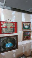 Four vintage alcohol signs displayed on a wall in a room with unfinished drywall. Signs include Budweiser, Olympia Beer, Heineken, and Geyser Peak Winery. Signs appear framed or bordered with wood or metal.