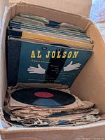 Box of multiple 78 RPM records showing Al Jolson record on top with blue sleeve and red label vinyl disc exposed.