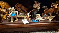 Display of multiple animal figurines including mountain lions, wolf, ram, clown, and eagle on wooden shelf showing scale and variety.