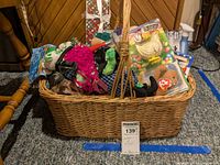 Wicker basket filled with assorted toys including plush Beanie Babies, packaged McDonald's toys, bobblehead figures, and toy vehicles, placed on carpeted floor against wood-paneled wall.