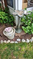 Both the cement bird bath and the planter with pink rose decoration shown together placed near garden plants.