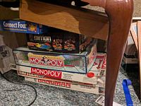 Stack of vintage 1980s board games under a table, showing Connect Four, Simon, Monopoly Deluxe Edition, Monopoly standard, Battleship, Hangman game boxes.