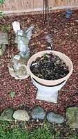 Outdoor garden scene with two concrete figurines (praying angel and person with flowers) next to a plastic planter on a concrete base, all placed in mulch surrounded by stones.