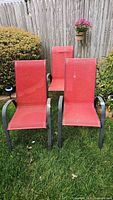 Three metal framed outdoor chairs with red fabric seats and backs shown outdoors on grass with wooden fence background.