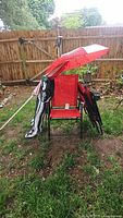 Photo of three red metal framed folding chairs, two folded black and white camping chairs, and a red two-piece umbrella outside on grass near a wooden fence.
