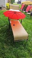 Full view of the lounge chair outdoors, featuring beige fabric on metal frame with an open red umbrella for scale and context.