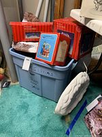 Boxes of Christmas village accessories in red packaging stacked on a gray plastic storage bin labeled with holiday contents. Items include skating rink, mountain, and river sets.