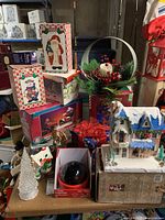 Photo showing boxed Santa and snowman decorations, projection light in original packaging, white Christmas tree figurine, winter village house with blue roof and snow accents, and various other holiday decorative items including a basket with pine and red berry decor.