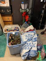 Photo showing plastic bins containing Christmas ornaments and garland, with plush snowflake blankets and some decorative items nearby.